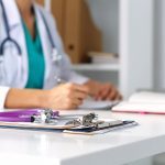 Stethoscope head lying on medical forms on clipboards closeup while medicine doctor working in background. Health care, insurance and help concept. Physician ready to examine patient